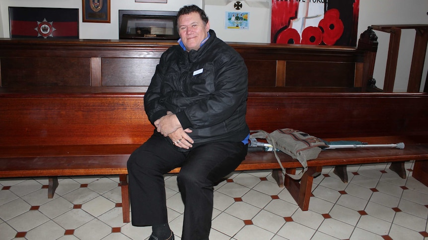 A man sits on a church pew with his hands folded in his lap, looking at the camera.