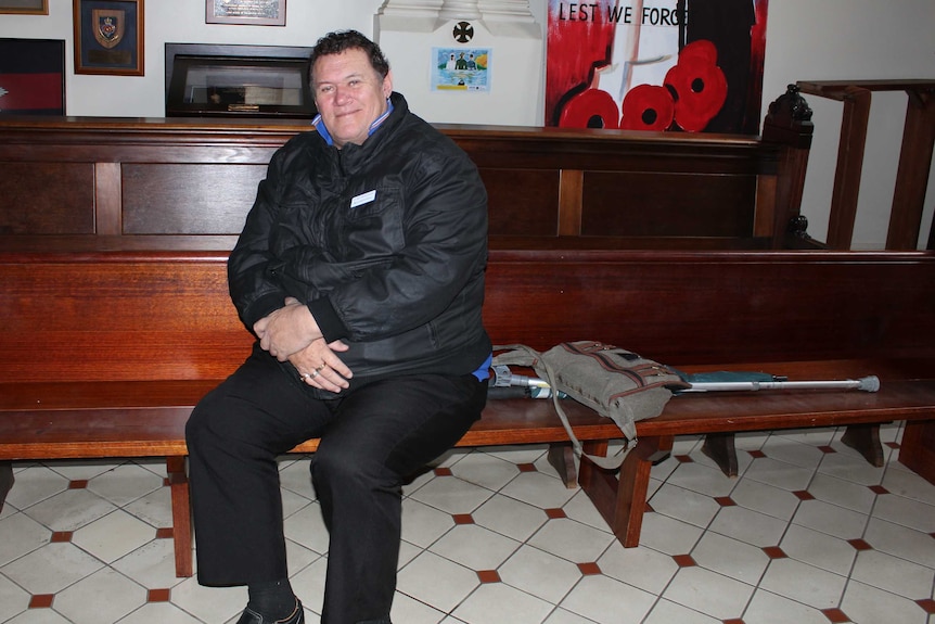 A man sits on a church pew with his hands folded in his lap, looking at the camera.