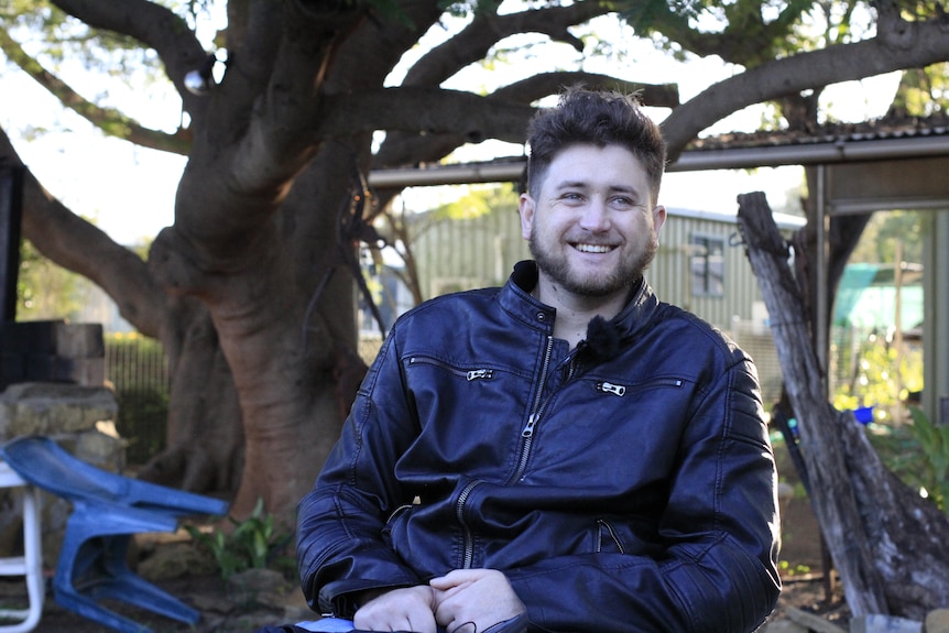 A young man in a wheelchair sits in the shade of some trees in a backyard.