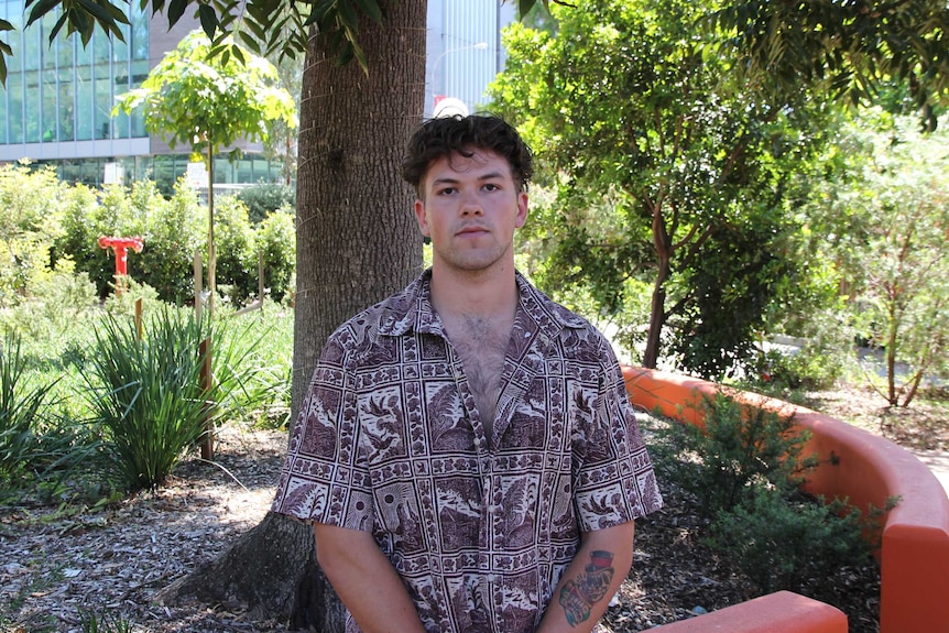 A young man stands under a tree with a serious expression on his face.