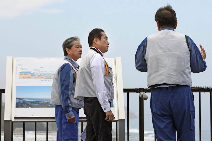 Three older Japanese men wearing grey vests look over a railing at the ocean.