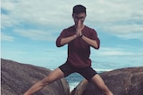 Man doing a yoga pose standing on a rock with the ocean behind him.