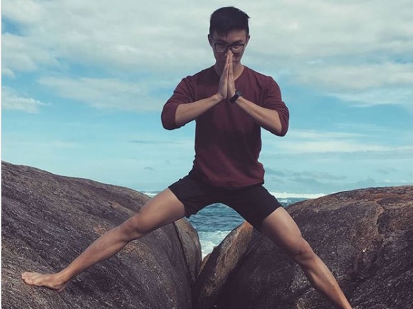Man doing a yoga pose standing on a rock with the ocean behind him.