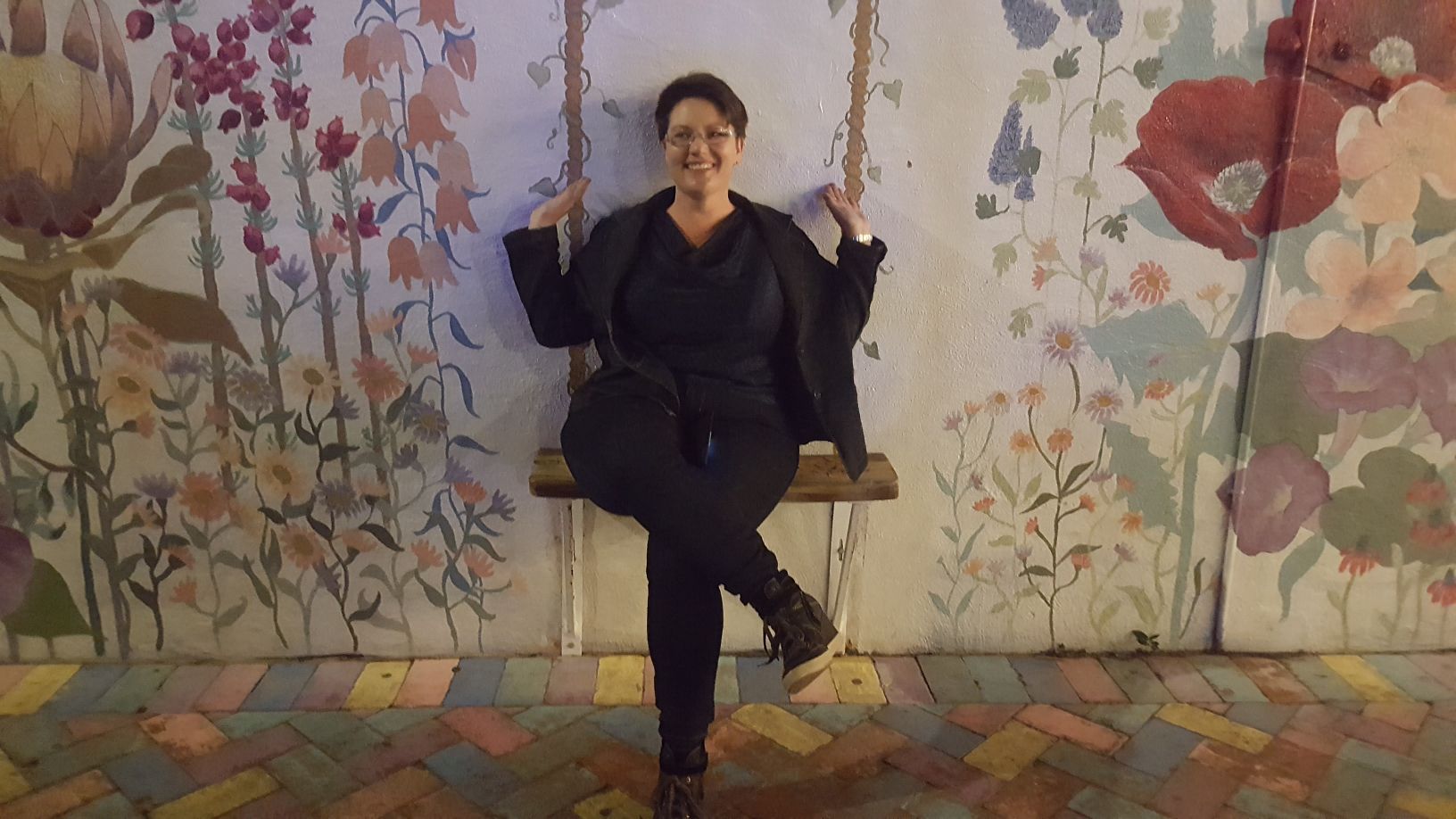 Улыбающаяся молодая женщина с короткими черными волосами и в очках, одетая в черное, сидит на стуле перед стеной с произведениями искусства.