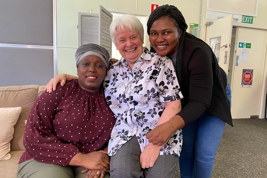 Three smiling women sitting side by side on a sofa indoors embracing each other