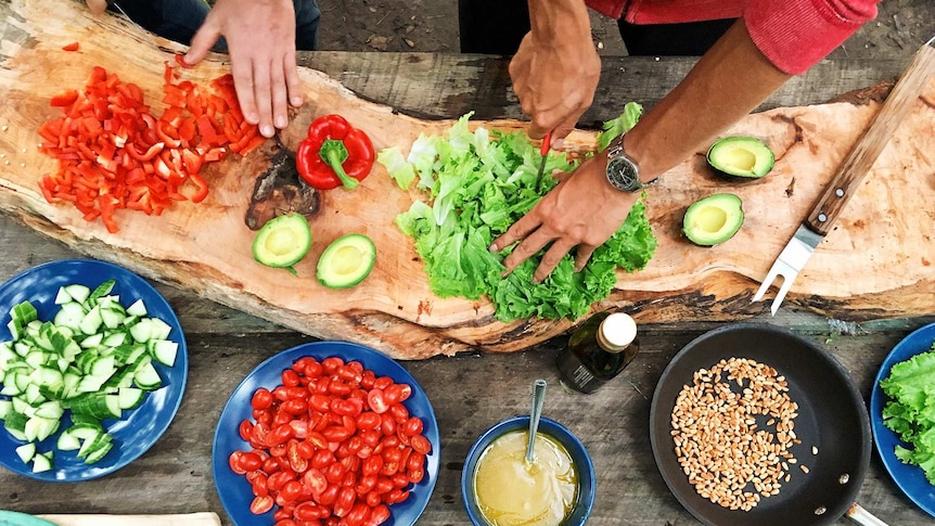 Overhead photo of two people cutting vegetables on a table with bowls of salad ingredients.