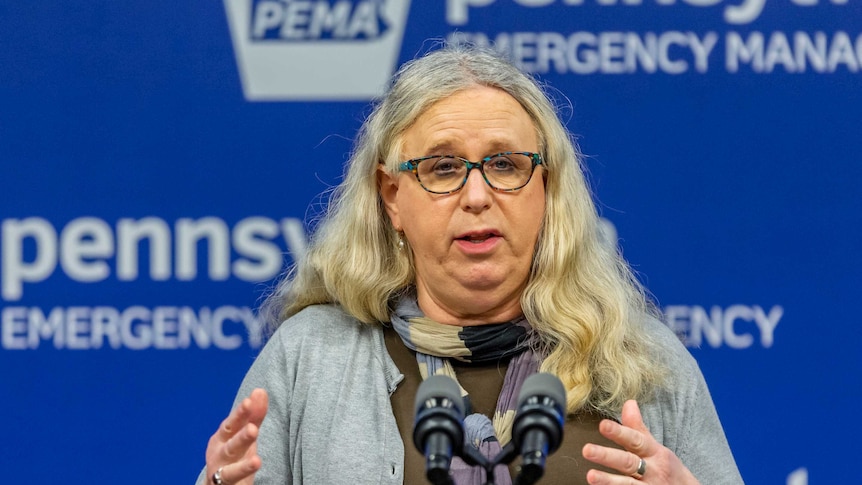 Woman with long greying hair and green glasses speaks at a microphone