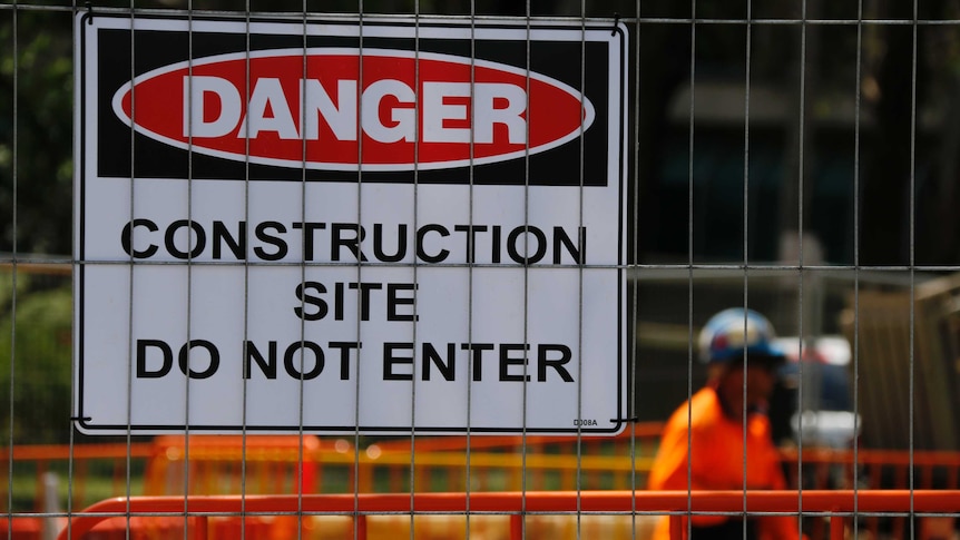 A sign that reads "DANGER Construction site, do not enter" is pinned to a fence. A worker in high vis is in the background.