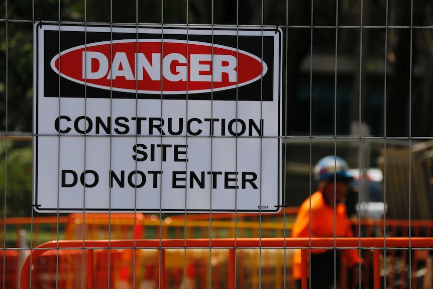 A sign that reads "DANGER Construction site, do not enter" is pinned to a fence. A worker in high vis is in the background.