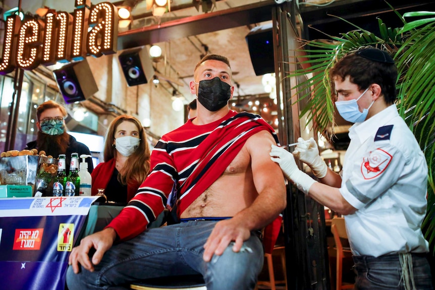 A masked man with his shirt half off in a bar sits while a paramedic injects his arm and a woman in the background watches on