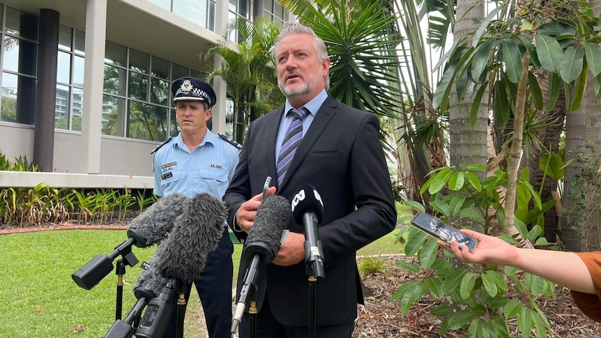A uniformed policeman stands behind a grey-haired, bearded man in a suit. They are on a lawn, addressing the media.