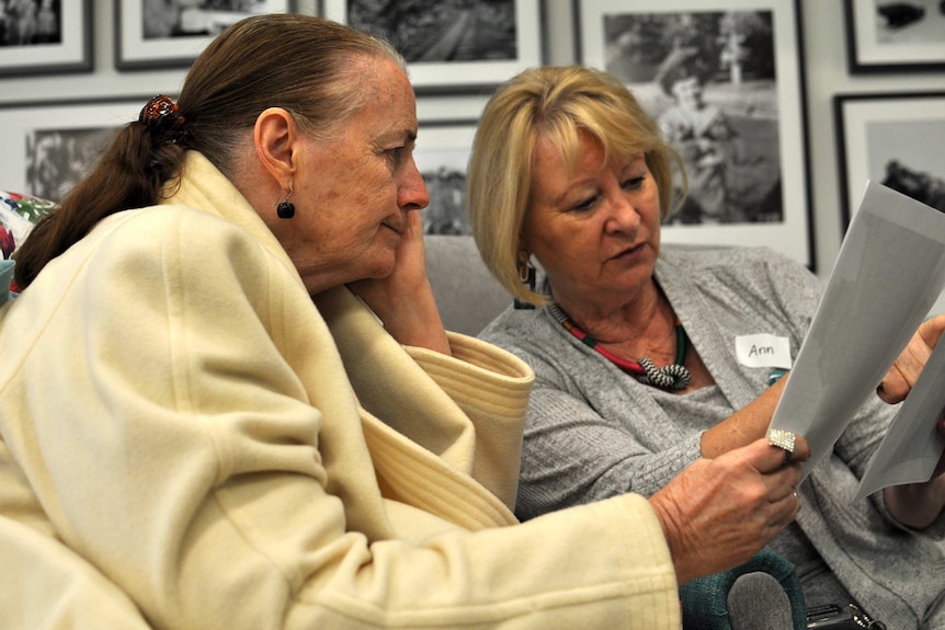 A woman holds her face while looking at a photo another woman is showing her.