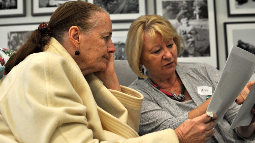 A woman holds her face while looking at a photo another woman is showing her.