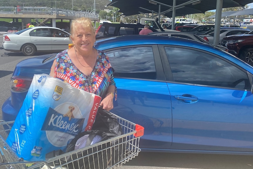 Carol Scott stands in front of a blue car, leaning on a trolley with a 32 pack of toilet paper.