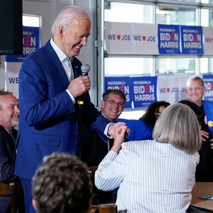 Joe Biden speaks into a microphone and holds the hand of a woman seated at a restaurant table.
