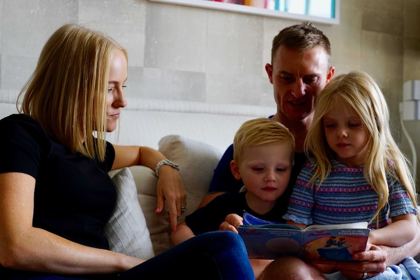 Zac Purton, Nicole Purton, and their children Roxy and Cash sitting on a sofa reading a children's book.