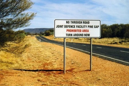A warning sign next to a road says "No through road. Joint defence facility Pine Gap. Turn around now."