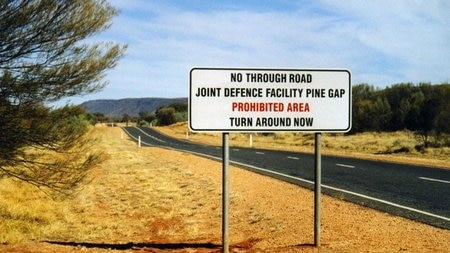 A warning sign next to a road says "No through road. Joint defence facility Pine Gap. Turn around now."