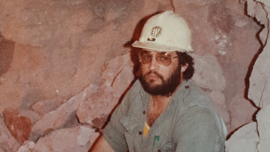 A bearded man in jeans, a miners hat sits casually in what appears to be a dry cave. He's wearing sunglasses and looking cool.