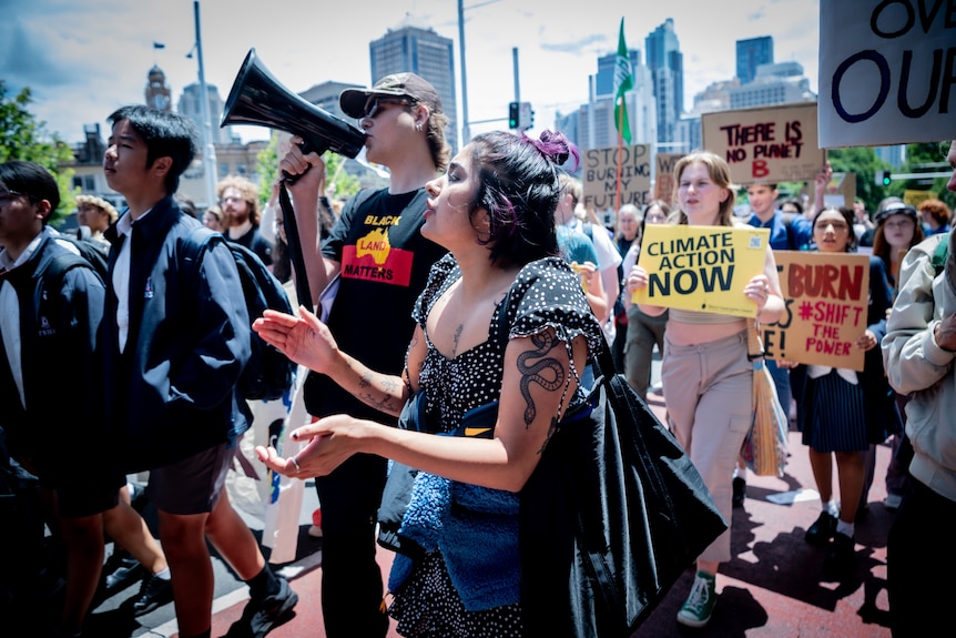 A girl a snake tattoo on her arm claps hands and chants, a man with a megaphone next to her, students marching behind.