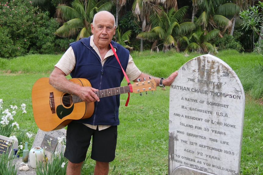 An older man stands with a guitar, next to a grave headstone from the Thompson family.