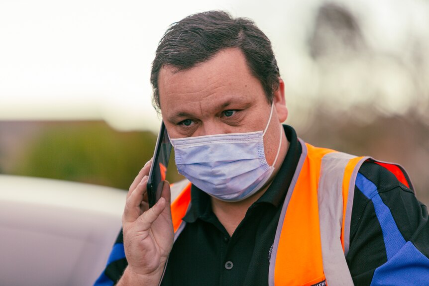 A man in an orange high-vis vest and surgical mask speaks on his mobile phone.