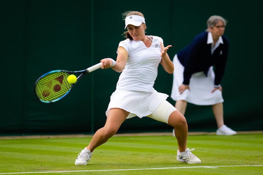 A white-clad female tennis player bends her wrist to hit a low forehand return at Wimbledon, a lineswoman in the background.