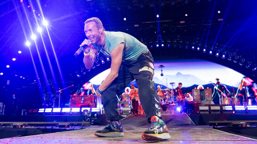 Chris Martin from Coldplay on stage singing with a microphone in hand and the band behind him