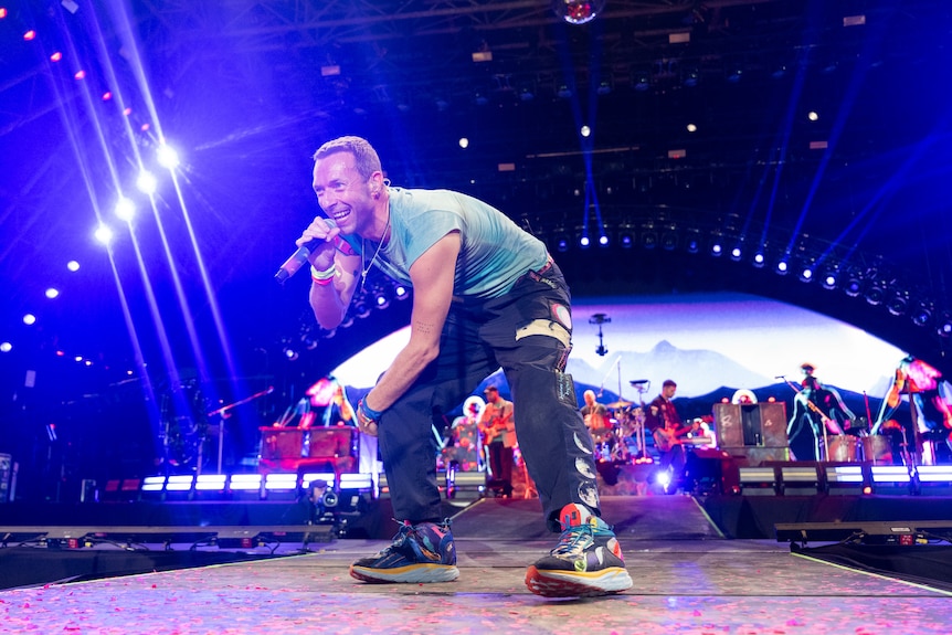 Chris Martin from Coldplay on stage singing with a microphone in hand and the band behind him
