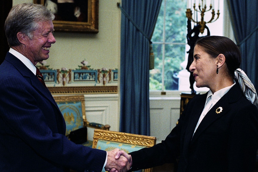 Colour photo of former President Jimmy Carter smiling and shaking hands with Ruth Bader Ginsburg in the oval office in 1980.