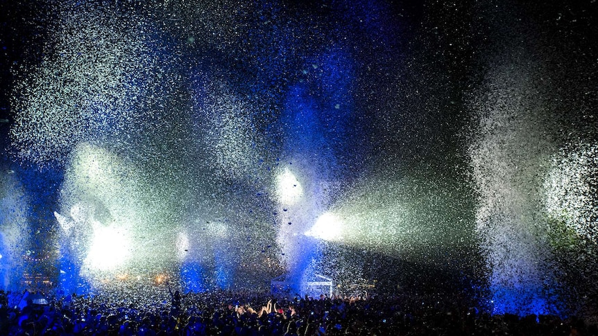Blue and white glitter fly through the air, illuminated by beams of light coming from a large stage.