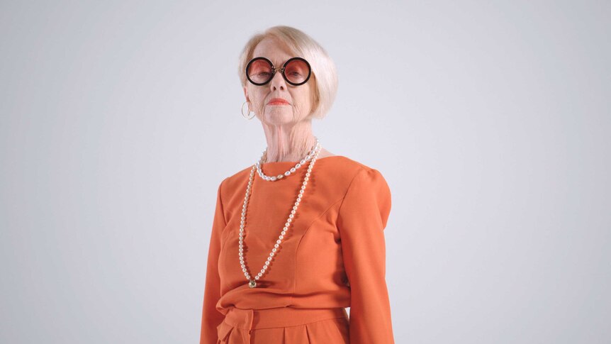 Older woman with white-blonde bob wearing orange jumpsuit, black-rimmed glasses and string of pearls, staring at camera.