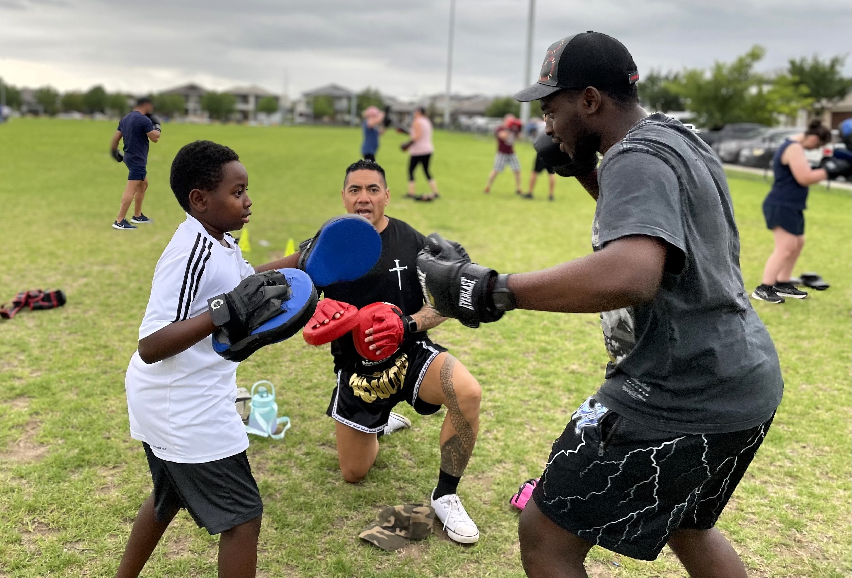A young boy and his older brother are sparring boxing, a man is crouched in the background and shouts instructions.