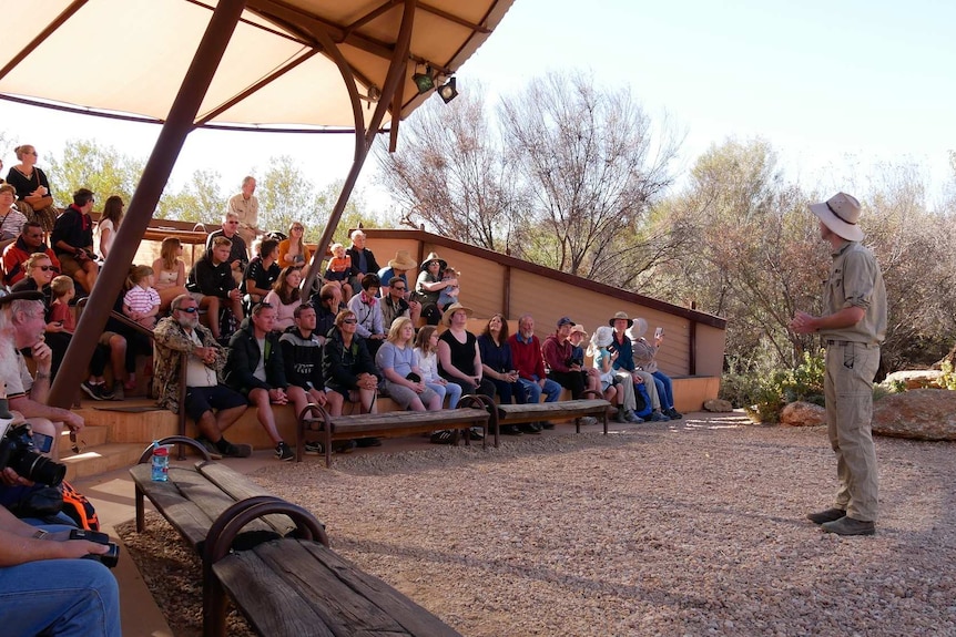 A man in khaki stands and speaks to bird show attendees  in the amphitheatre at the Alice Springs Desert Park