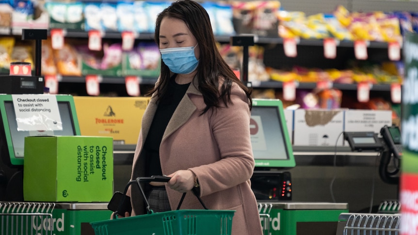 a woman wearing a mask carrying a shopping basket inside a grocery store