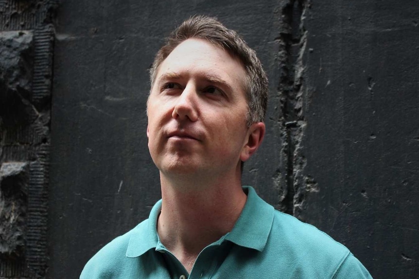 Tim Hillier, a man in his 30s wearing a green shirt, looks up to the right in front of a black wall.