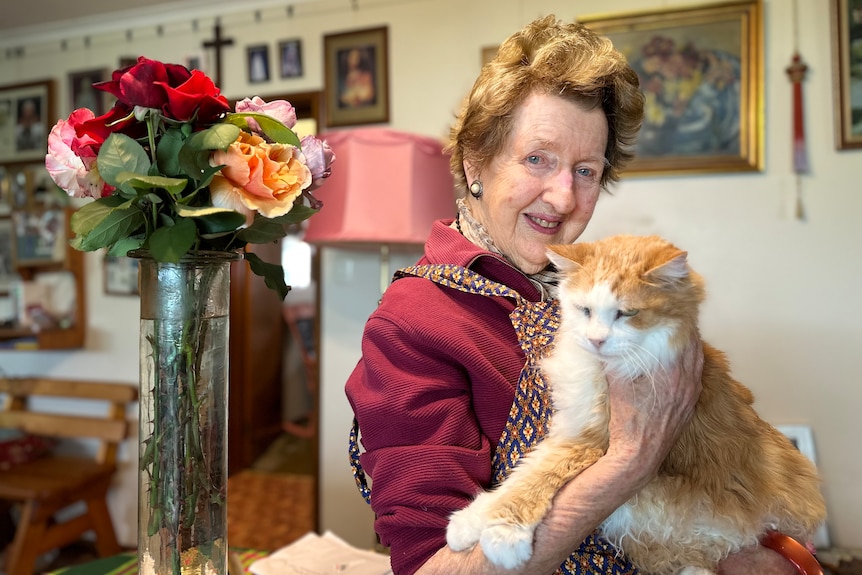 An elderly woman with her hair swept up, holds a ginger and white cat while standing next to a vase of roses.