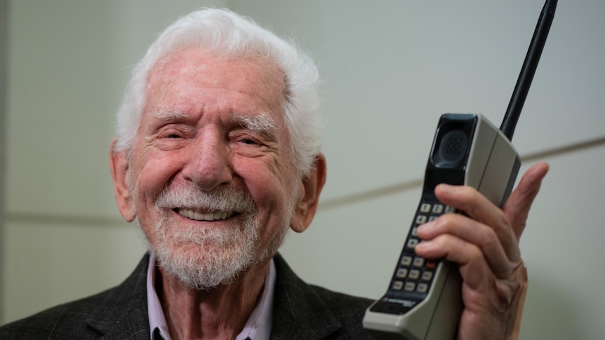 An elderly man poses for a photo holding one of the first commercial mobile phones