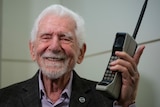 An elderly man poses for a photo holding one of the first commercial mobile phones
