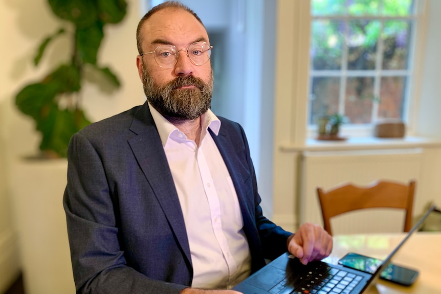 A man in a suit looking serious and sitting at a dining room table, with a laptop in front of him.