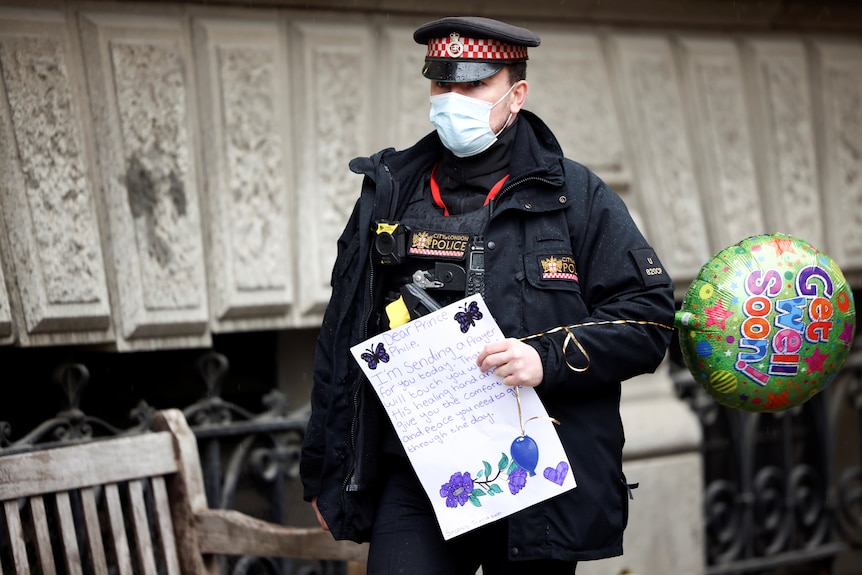 A police officer carries a balloon reading 'get well soon' and a handwritten note praying for Prince Philip's recovery.