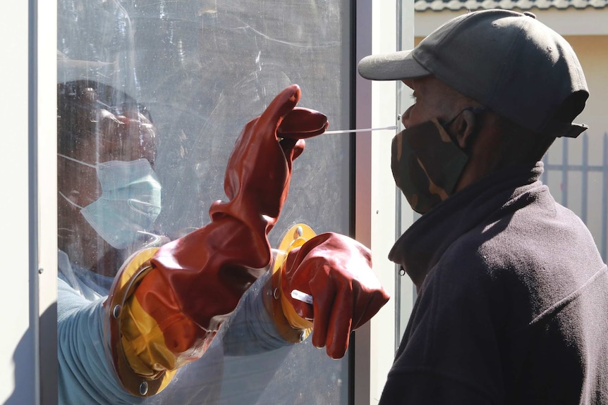 A health worker stands behind a clear screen wearing PPE as she uses a long white swab to take a nasal sample from a man