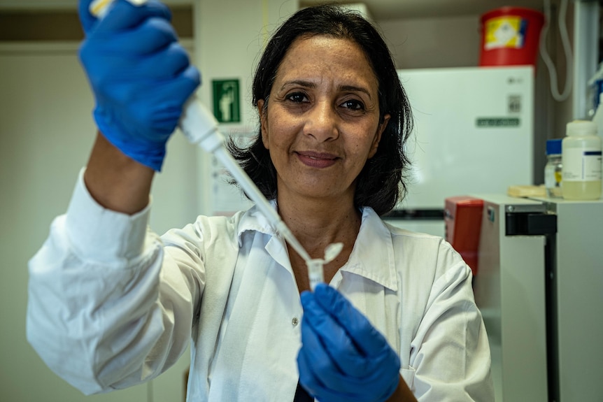 A woman in a white lab coat holds up a test tube while wearing blue gloves 