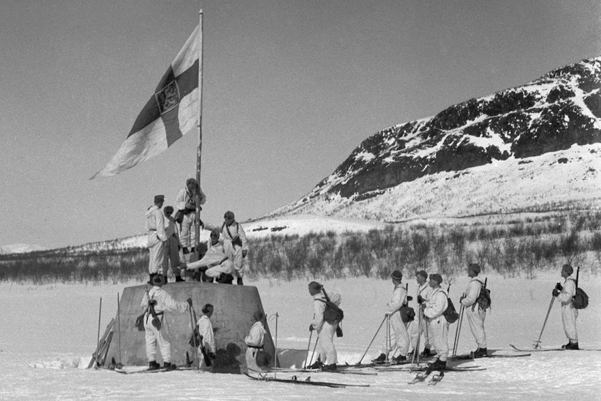 On an icy field with mountains in the distance, a group of Finnish soldiers on skis raise a Finnish flag on a concrete mount.