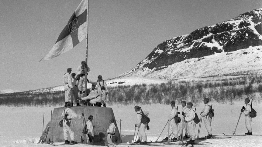 On an icy field with mountains in the distance, a group of Finnish soldiers on skis raise a Finnish flag on a concrete mount.