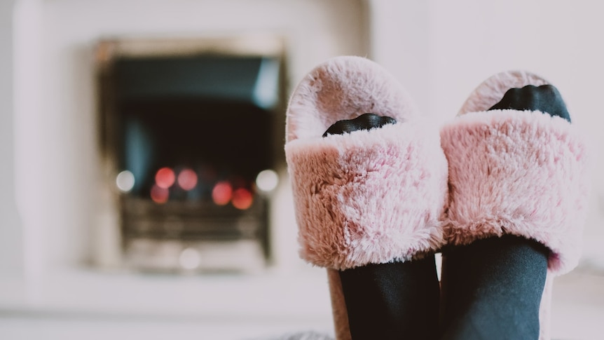 A close up of two feet wearing pink fluffy slippers. Out of focus in the background is a lit fireplace.