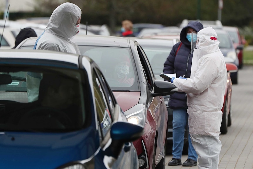 Two people in PPE stand around a car as one of them examines some paperwork