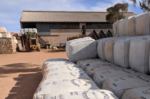 Big white bales of packaged wool sit on top of each other outside shearing shed