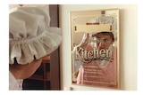 A woman in a white frilly har checks her reflection in a mirrored sign reading 'Gourmet Kitchen'