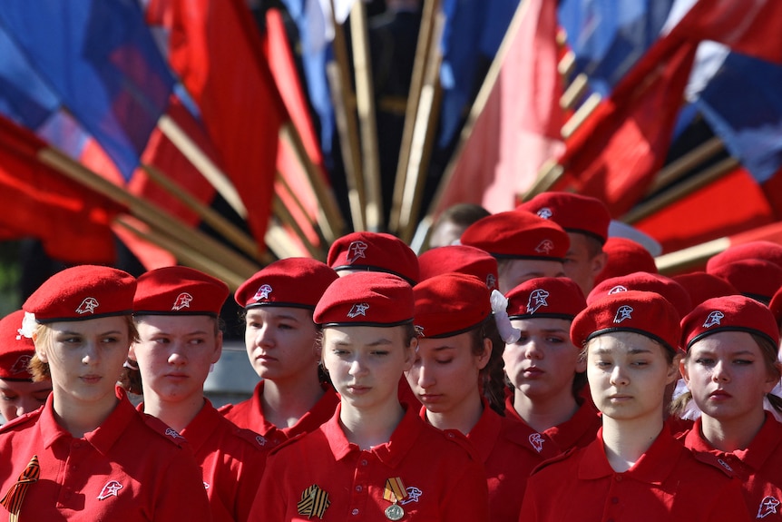 A group of young people dressed in red uniforms and hats stand in front of Russian flags.